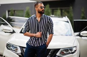 succesvolle arabische man draagt een gestreept shirt en een zonnebril poseert in de buurt van zijn witte suv-auto. stijlvolle arabische mannen in transport. foto