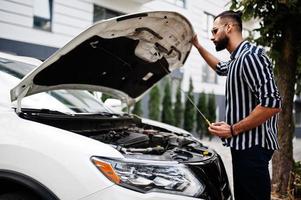 succesvolle arabische man draagt een gestreept shirt en een zonnebril poseert in de buurt van zijn witte suv-auto, controleer de motor met open kap. foto