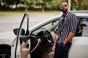 succesvolle arabische man draagt een gestreept shirt en een zonnebril poseert in de buurt van zijn witte suv-auto. stijlvolle arabische mannen in transport. foto