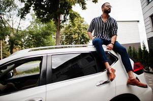 succesvolle arabische man draagt een gestreept shirt en een zonnebril die op het dak van zijn witte suv-auto zit. foto