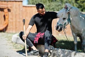 Arabische hipster baard man houthakker houd bijl tegen pony paard. foto