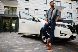 succesvolle arabische man draagt een gestreept shirt en een zonnebril poseert in de buurt van zijn witte suv-auto. stijlvolle arabische mannen in transport. foto