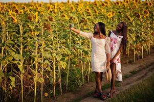 twee mooie jonge zwarte vrienden vrouw dragen zomerjurk poseren in een zonnebloemveld selfie maken op de telefoon. foto