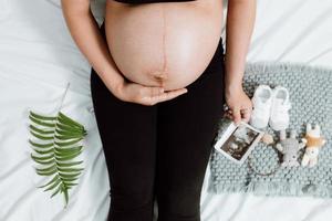 zwangere vrouwenhanden die op buik houden maken een zorg en liefdesymbool op de slaapkamer met ultrasone klankbeeld. aanstaande moeder die wacht en zich voorbereidt op de geboorte van een baby. zwangere vrouw concept. foto
