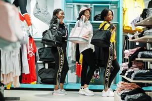 drie Afro-Amerikaanse vrouwen in trainingspakken winkelen met sporttassen in sportkledingwinkelcentrum tegen planken. sport winkel thema.