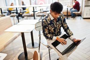 jonge mannelijke Indiase freelancer zit op een opengeklapte laptopcomputer in een fastfoodcafé, een knappe aziatische man die een sms leest tijdens het werk aan een netbook in een comfortabele coffeeshop. foto