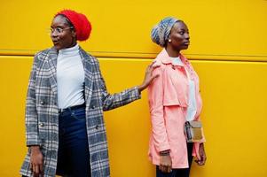 twee jonge moderne modieuze, aantrekkelijke, lange en slanke Afrikaanse moslimvrouwen in hijab of tulband hoofddoek en jas gesteld tegen gele bus. foto