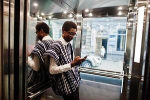 afrikaanse man in traditionele kleding en bril met mobiele telefoon op de lift of moderne lift. foto