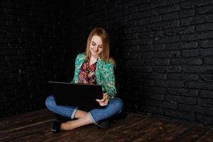 stijlvol blond meisje in jas en spijkerbroek met laptop tegen zwarte bakstenen muur in de studio. foto