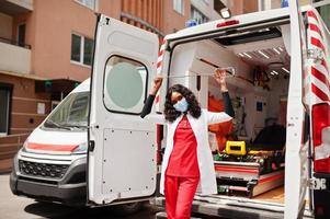 Afro-Amerikaanse vrouwelijke paramedicus in gezichtsbeschermend medisch masker dat voor de ambulanceauto staat. foto