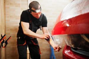 autoservicemedewerker plaatste anti-grindfilm op een rode carrosserie in de detailleringsvoertuigwerkplaats. autobescherming met speciale folies. foto