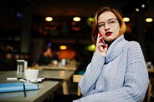 vrolijke jonge mooie roodharige vrouw in glazen zittend op haar werkplek op café en spreken via mobiele telefoon. foto