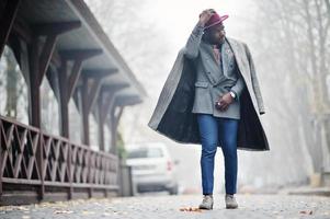 stijlvolle Afro-Amerikaanse man model in grijze jas, jas stropdas en rode hoed gesteld op mistig weer straat. foto