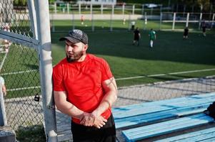 jonge brute bebaarde gespierde man draagt een rood shirt, korte broek en pet in het stadion. foto