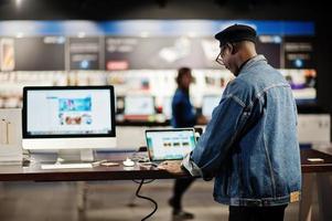 stijlvolle casual Afro-Amerikaanse man bij jeans jasje en zwarte baret met behulp van nieuwe laptop bij elektronicawinkel. foto