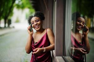 portret van een mooie natuurlijke jonge afrikaanse vrouw met afrohaar. zwart model in rode zijden jurk met mobiele telefoon. foto