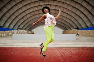 geweldige Afro-Amerikaanse modelvrouw in groene broek en zwarte hoed poseerde buiten tegen de arenahal, spring in de lucht. foto