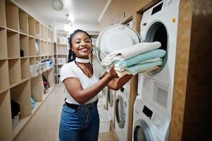 vrolijke Afro-Amerikaanse vrouw met handdoeken in handen in de buurt van wasmachine in de zelfbedieningswasserette. foto