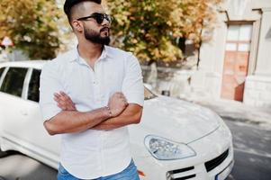 stijlvolle lange Arabische man model in wit overhemd, jeans en zonnebril gesteld op straat van de stad. baard rijke aantrekkelijke arabische man tegen witte suv auto. foto