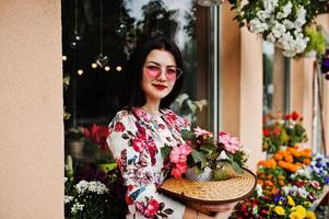 zomer portret van brunette meisje in roze bril en hoed tegen bloemenwinkel. foto