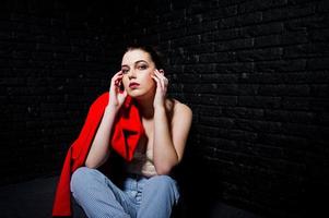 stijlvolle brunette meisje op rode jas tegen studio zwarte bakstenen muur. foto