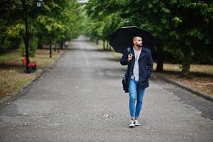 modieuze lange Arabische baard man draagt zwarte jas met paraplu en tas geval gesteld op regenweer dag. foto