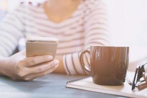 close-up mensen vrouw zitten met behulp van mobiele smartphone in een pauze ontspannen. kopje koffie op bureau tafel met boek notitieblok, brillen, apparatuur leveringen in het werk. ruimte kopiëren. concept werkende vrouw. foto