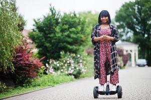 mooie afrikaanse amerikaanse vrouw die segway of hoverboard gebruikt. zwarte meid op zelfbalancerende elektrische scooter met twee wielen met mobiele telefoon bij de hand. foto