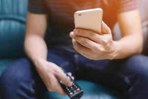 jonge man met behulp van de afstandsbediening van de televisie en mobiele smartphone zit op een bank. aan of uit tv. foto