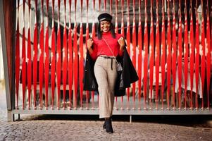 Afro-Amerikaanse mode meisje in jas en krantenverkoper GLB gesteld op straat tegen rode achtergrond. foto