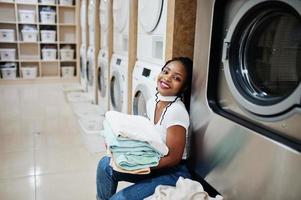 vrolijke Afro-Amerikaanse vrouw met handdoeken in handen in de buurt van wasmachine in de zelfbedieningswasserette. foto
