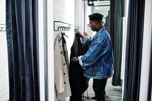 stijlvolle casual Afro-Amerikaanse man bij jeans jasje en zwarte baret bij paskamer kleding winkel wissel overjas. foto
