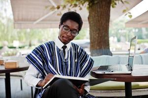 afrikaanse man in traditionele kleding en bril die achter de laptop zit in een buitencafé en op zijn notitieboekje kijkt.