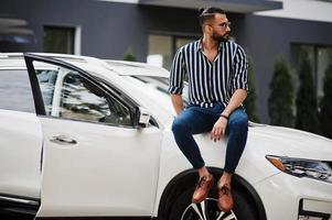 succesvolle arabische man draagt een gestreept shirt en een zonnebril die op de motorkap van een witte suv-auto zit. stijlvolle arabische mannen in transport. foto