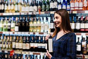 winkelende vrouw die naar de schappen in de supermarkt kijkt. portret van een jong meisje in een marktwinkel in de alcoholsector met een fles wijn bij de hand. foto