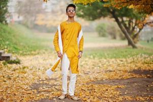 Indiase stijlvolle man in gele traditionele kleding met witte sjaal, zonnebril poseerde buiten tegen de boom van de herfstbladeren. foto