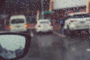 druppels regen motregen op de glazen voorruit in de avond. straat in de zware regen. bokeh achterlicht en verkeerslichten in de stad. rijd alstublieft voorzichtig met de auto, gladde weg. zachte focus. foto