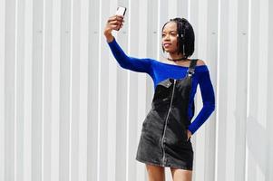 afrikaanse vrouw in denim overalls rok en blauwe jurk poseerde tegen een witte stalen muur en maakte selfie op de telefoon. foto