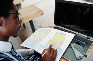 afrikaanse man in traditionele kleding en bril die achter een laptop zit in een buitencafé en op de kaart van afrika en ghana naar zijn notitieboekje kijkt. foto