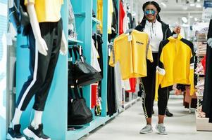 Afro-Amerikaanse vrouwen in trainingspakken en zonnebrillen winkelen bij sportkleding winkelcentrum tegen planken. ze koos geel t-shirt. sport winkel thema. foto