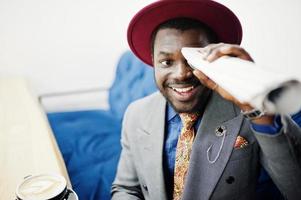 stijlvolle Afro-Amerikaanse man model in grijze jas stropdas en rode hoed koffie drinken in café en met kranten buis. foto