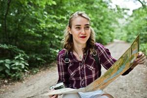 portret van een positieve jonge prachtige blonde zittend op de grond met een kaart in haar handen in het bos. foto