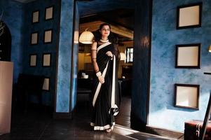jonge indiase vrouw draagt een elegante zwarte saree die op restaurant tegen de muur met frames staat. foto