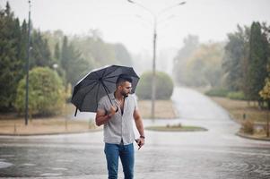 modieuze lange Arabische baard man slijtage op shirt, jeans en zonnebril met paraplu gesteld bij regen op parkplein. foto