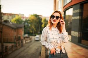 aantrekkelijke roodharige vrouw in zonnebril, draag op witte blouse poseren op straat tegen modern gebouw. foto