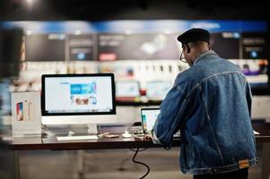 stijlvolle casual Afro-Amerikaanse man bij jeans jasje en zwarte baret met behulp van nieuwe laptop bij elektronicawinkel. foto