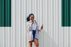 afrikaanse vrouw met dreadlocks haar, in spijkerbroek gesteld tegen witte stalen muur met mobiele telefoon in de hand. foto