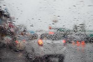 druppels regen motregen op de glazen voorruit in de avond. straat in de zware regen. bokeh achterlicht en verkeerslichten in de stad. rijd alstublieft voorzichtig met de auto, gladde weg. zachte focus. foto