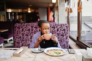 Afrikaanse vrouw in geruite violette cape en bril poseerde in café, zittend aan tafel met dessert en kopje koffie. foto