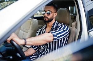 succesvolle arabische man draagt een gestreept shirt en een zonnebril poseert achter het stuur van zijn witte suv-auto. stijlvolle arabische mannen in transport. foto